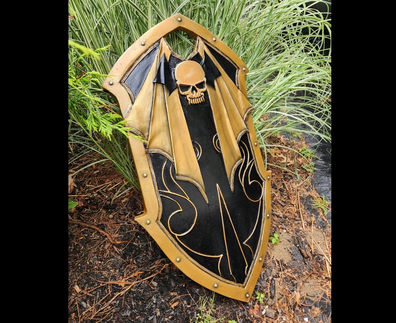 K&ouml;nnte beinhalten: Ein schwarzer und goldener Schild mit einem Totenkopf- und Fl&uuml;gelmotiv. Der Schild ist aus Holz und hat eine abgenutzte Oberfl&auml;che.