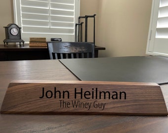 May include: A dark brown wooden desk nameplate with the text "John Heilman The Winey Guy" engraved in black.