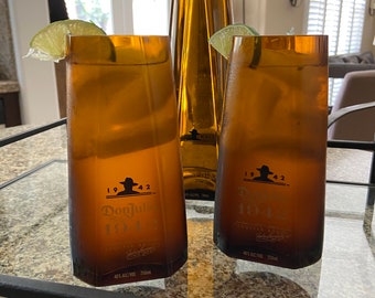 May include: Two amber-colored glasses filled with a clear liquid and garnished with lime wedges. The glasses are on a glass surface with a black metal frame. The glasses have labels that read "Don Julio 1942 Tequila" and "Don Julio 1944 Tequila".