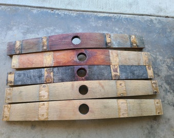 May include: Five reclaimed wooden barrel staves with varying colors, including brown, gray, and tan. Each stave has a circular hole and metal bands. The staves are arranged horizontally, showcasing their unique wood grain.