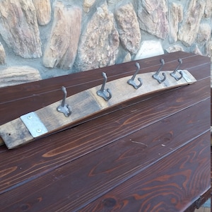 May include: A rustic wooden coat rack made from a reclaimed wine barrel with five metal hooks. The rack is mounted on a dark brown wooden wall.