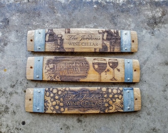 May include: Three wooden wine barrel stave signs with metal accents. Each sign features a different design: a wine bottle and glass, a wine glass, and a bunch of grapes. The text on each sign reads "The Jordans Wine Cellar".
