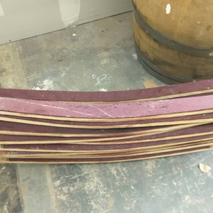 May include: A stack of curved wooden barrel staves with a reddish-brown color. The staves are stacked on top of each other and have a slight curve to them. The staves are made of wood and have a natural finish.