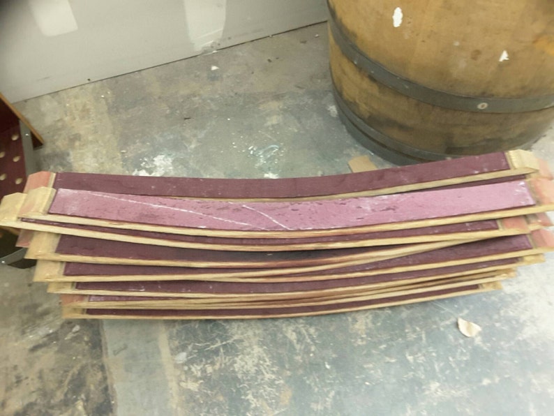 May include: A stack of curved wooden barrel staves with a reddish-brown color. The staves are stacked on top of each other and have a slight curve to them. The staves are made of wood and have a natural finish.