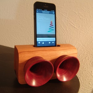 Pode incluir: Um dock de alto-falante de madeira com dois alto-falantes grandes de cor vermelha escura. Um smartphone preto está no dock, reproduzindo música do aplicativo Pandora. A tela mostra a capa do álbum "Shades of Christmas" de The Holly and The Ivy.