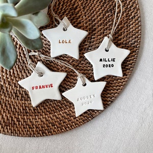 May include: Four white star-shaped decorations, each with a name and date. The names Lola, Frankie, Millie, and Audrey are written on the stars in gold, red, and black. Each decoration has a string for hanging. They are on a woven surface.