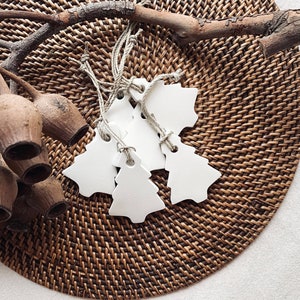 May include: Several white, Christmas tree-shaped gift tags with twine, arranged on a woven brown placemat. Dried eucalyptus pods and a branch add to the natural, rustic feel. The tags are a simple, elegant design, perfect for festive gifting.