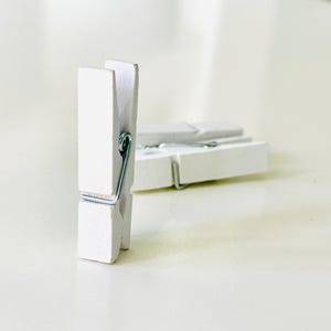 Puede incluir: Tres pinzas de ropa de madera blanca con muelles de metal plateado. Una pinza está de pie, mientras que las otras dos están tumbadas. Las pinzas de ropa están sobre una superficie de color claro.