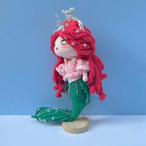 Op de afbeelding: Een gehaakte zeemeerminpop met rood haar, een roze top en een groene staart. De pop draagt een zilveren kroon met witte kralen en staat op een houten voet.