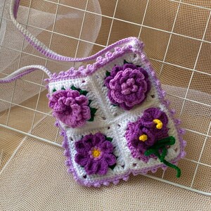 May include: A crocheted purple and white floral bag with a long strap. The bag is made of squares with different flower designs.