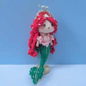 Op de afbeelding: Een gehaakte zeemeerminpop met rood haar, een roze top en een groene staart. De pop draagt een kroon met witte kralen en staat op een houten voet.