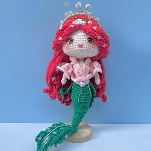 Op de afbeelding: Een gehaakte zeemeerminpop met rood haar, een roze top en een groene staart. De pop draagt een kroon met strass en staat op een houten voet.