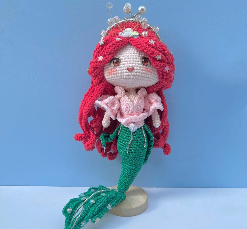 Op de afbeelding: Een gehaakte zeemeerminpop met rood haar, een roze top en een groene staart. De pop draagt een kroon met strass en staat op een houten voet.