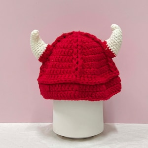 Puede incluir: Sombrero rojo de ganchillo con cuernos blancos. El sombrero está sobre una superficie blanca.