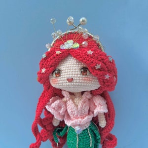 Op de afbeelding: Een gehaakte pop met rood haar, een roze top en een groene rok. De pop draagt een kroon met zilveren en witte kralen.