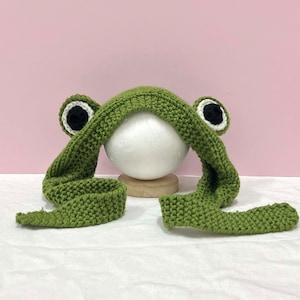 May include: Green knitted frog hat with black eyes and a long tail. The hat is on a white surface.