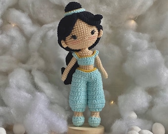 May include: A crocheted doll with dark hair, wearing a blue and yellow outfit. The doll is standing on a wooden base.