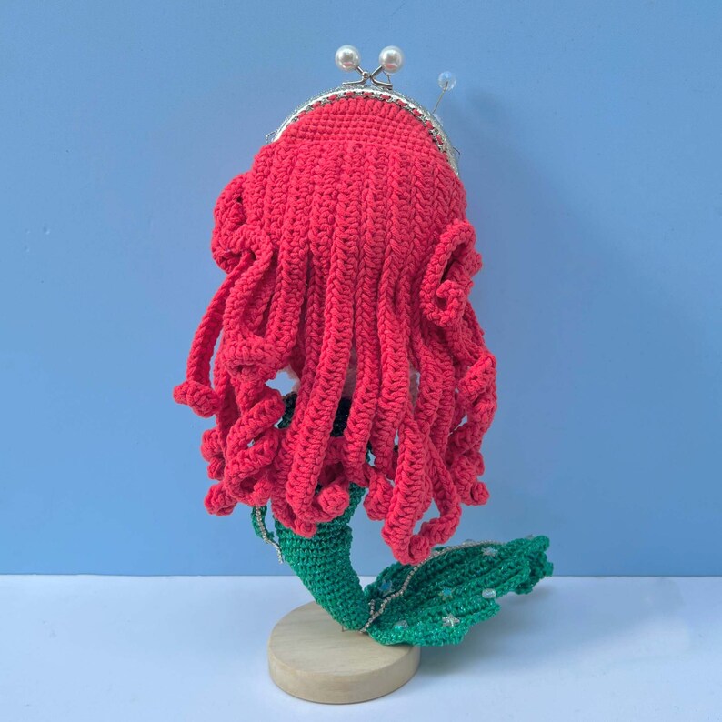 Op de afbeelding: Een gehaakte zeemeerminpop met rood haar en een groene staart. De pop draagt ​​een parelketting en heeft een gehaakte handtas.