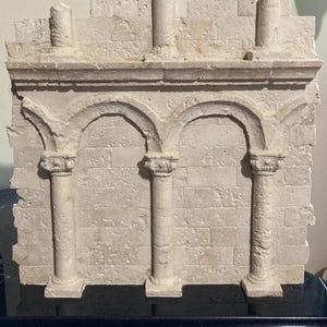 May include: A beige, rectangular architectural relief sculpture featuring two arched openings supported by columns. The sculpture has a textured, aged appearance, with a brick-like pattern on the background and decorative elements.