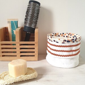 Peut inclure: Un organisateur en bois contient une brosse à cheveux ronde et un tube de produit. Un panier en tissu blanc avec un bord à motifs et une garniture décorative est à proximité. Un pain de savon repose sur un porte-savon en luffa naturel.