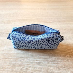 Peut inclure: Petite trousse ouverte, ornée d'un motif de cœurs bleus sur fond bleu clair. L'intérieur présente une doublure rayée bleue et blanche et une étiquette en cuir marron avec le texte "JoHCo". La fermeture éclair est bleue.