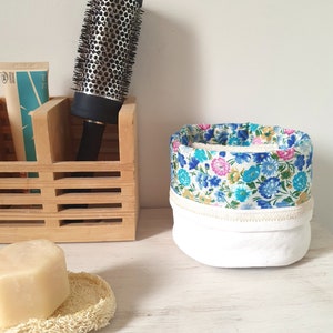 Peut inclure: Une brosse à cheveux ronde avec un manche noir et un barillet en métal est dans un organisateur en bois. Un panier en tissu floral avec des fleurs bleues, roses et jaunes est à côté. Un pain de savon repose sur un luffa.