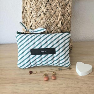 May include: A small, rectangular pouch with a green and white striped pattern. The pouch has a black label with the text "JoHCo". A small bow is attached to the zipper. The pouch is on a wooden surface with jewelry and a heart-shaped object.