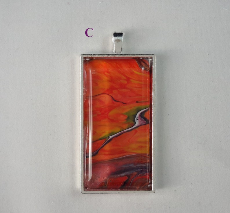 May include: A silver pendant with a rectangular glass insert featuring abstract red, orange, and yellow paint swirls.