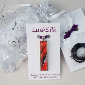 May include: A red, black, and white fluid acrylic painting on a silver pendant with a black cord necklace. The pendant is displayed on a white card with the text "LushSilk" and "Original Fluid Acrylic Painting" with an email address. The necklace is packaged in a white bag with a purple ribbon.