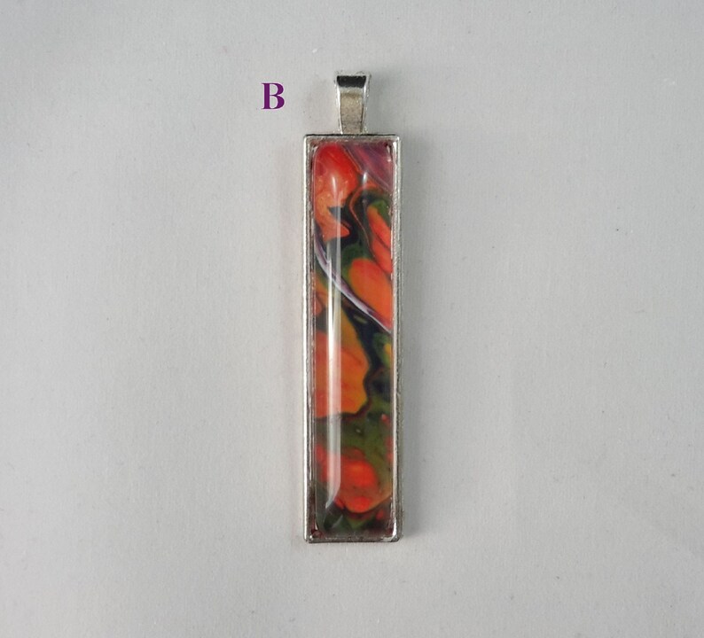 May include: A silver pendant with a rectangular glass insert featuring abstract swirls of orange, green, and black paint.