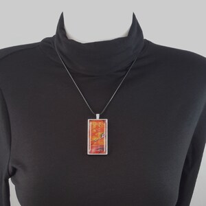 May include: A long, rectangular pendant necklace with a black cord. The pendant is made of clear resin with a red and orange abstract design.