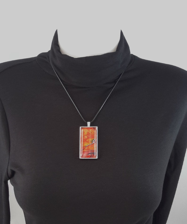 May include: A long, rectangular pendant necklace with a black cord. The pendant is made of clear resin with a red and orange abstract design.