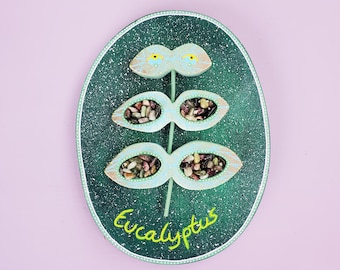 May include: A green oval-shaped wooden wall hanging with a stylised eucalyptus leaf design. The leaf is filled with small colourful stones and has yellow eyes at the top. The text "Eucalyptus" is written in green at the bottom.