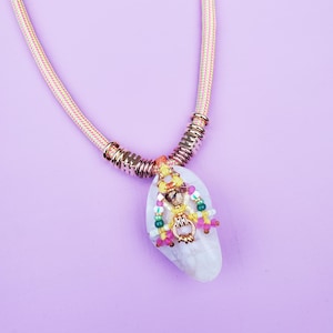 Peut inclure: Un collier artisanal avec un pendentif blanc de forme ovale orné de perles colorées et d'un charm doré. Le pendentif est suspendu à une chaîne en corde crème avec des accents dorés. Le collier est posé sur un fond violet.