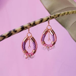 Peut inclure: Paire de boucles d'oreilles en forme de goutte d'eau, avec des détails tissés roses et violets, des accents métalliques dorés et de petites perles de cristal transparentes. Les boucles d'oreilles sont suspendues à des crochets dorés.