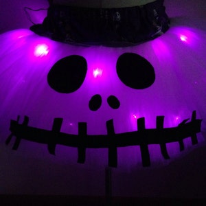 May include: A white tulle tutu skirt with a black skull and crossbones design. The skirt has purple LED lights that illuminate the design.