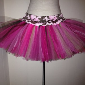 May include: A pink and brown tutu with a camouflage waistband. The tutu is made of multiple layers of tulle and has a sparkly finish.