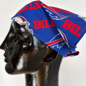 May include: A blue bandana with the words "BILLS" in red, along with the team logo. The bandana is displayed on a black mannequin head. The fabric has a repeating pattern of the team's logo.