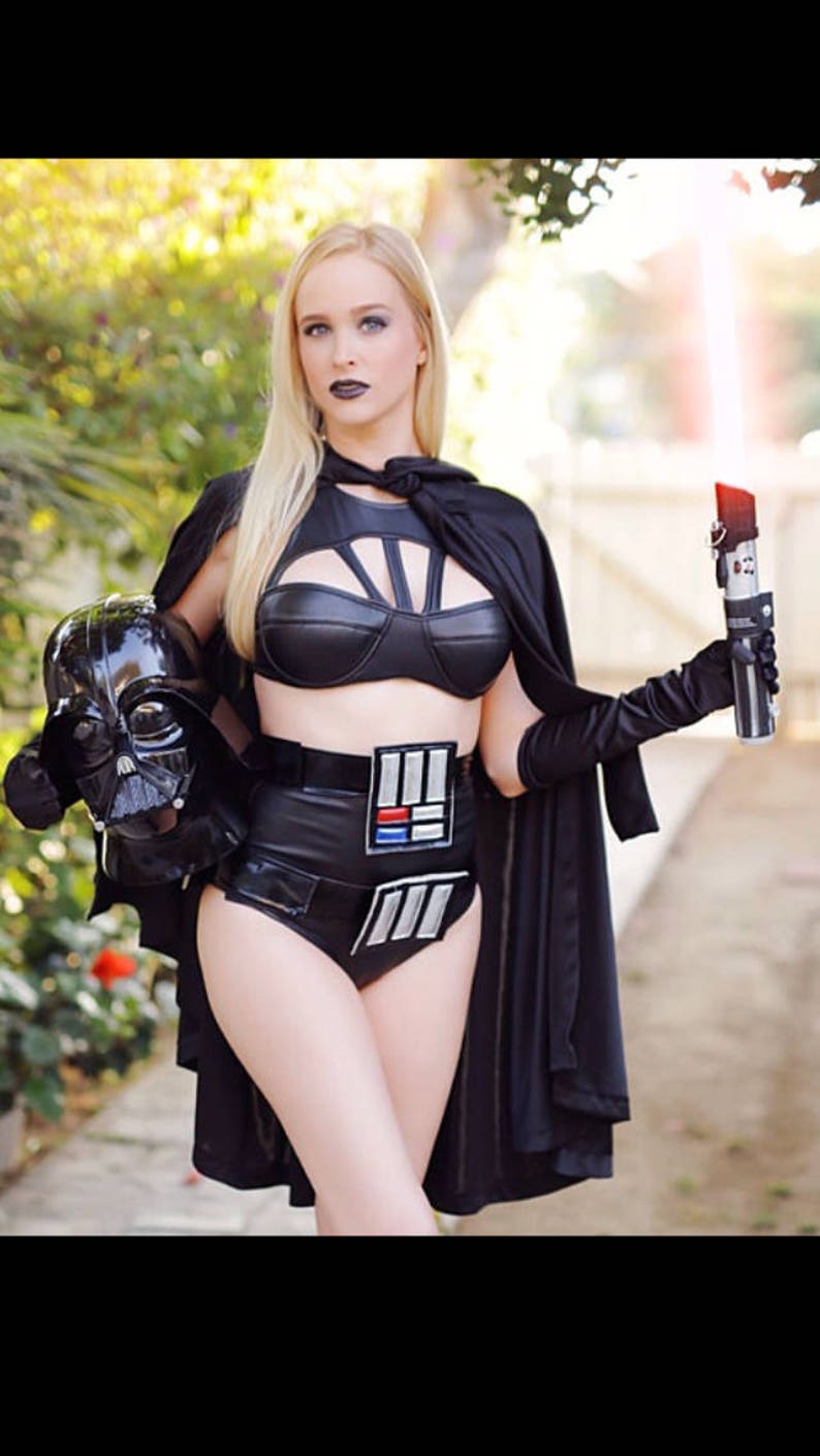May include: A woman dressed as Darth Vader from Star Wars. She is wearing a black leather bra and matching bikini bottoms with the Star Wars logo. She is holding a red lightsaber and a Darth Vader mask.