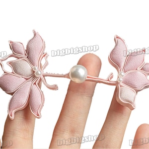 Puede incluir: Un par de pinzas para el pelo de flores de tela rosa con detalles de perlas. Las pinzas están conectadas por un alambre delgado con una perla en el centro.