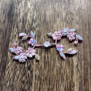 May include: A floral hair accessory with pink, blue, and white flowers and green leaves. The decorative piece is set against a wooden background. The flowers have a pearl-like accent.