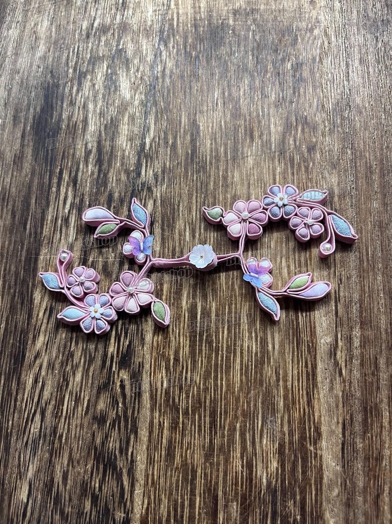 May include: A floral hair accessory with pink, blue, and white flowers and green leaves. The decorative piece is set against a wooden background. The flowers have a pearl-like accent.