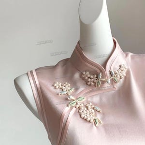 May include: A pale pink satin cheongsam dress with floral detailing. The sleeveless dress has a mandarin collar and decorative floral accents with pearl-like beads. The dress is displayed on a white mannequin.