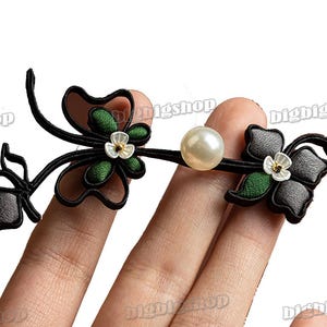 May include: A black and green fabric flower with a white pearl centre, attached to a black fabric stem with a second black and grey fabric flower. The flowers are on a hand.