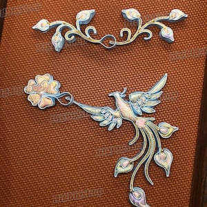 May include: Two decorative metal pieces with a floral and bird theme. The top piece is a floral vine with light blue and gold accents. The bottom piece features a bird with outstretched wings, flowers, and trailing leaves, in similar colors.