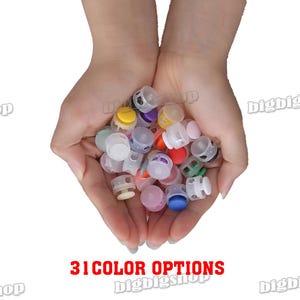 May include: A variety of colorful plastic cord locks in a person's hands. The cord locks are small and round, and they come in a variety of colors, including red, blue, green, yellow, and purple.