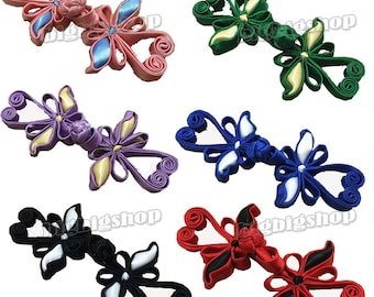 Op de afbeelding: Zes paar decoratieve Chinese knopen van stof in verschillende kleuren, waaronder roze, groen, paars, blauw, zwart en rood. Elke knoop heeft een vlinderontwerp met een bloem in het midden.