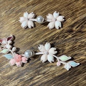 May include: Close-up of two sets of floral hair accessories. The accessories feature white and pink fabric flowers with pearl accents. One set is a pair of flowers connected by a pearl, and the other is a floral arrangement with leaves.
