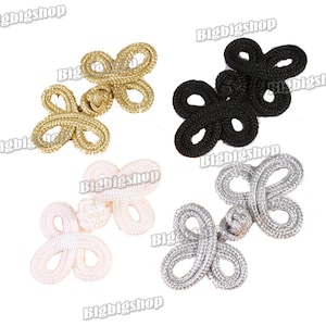 May include: Four decorative frog closures in gold, black, white, and silver. Each closure is made of a looped cord design, ideal for embellishing garments or crafts. The closures are displayed on a white background.