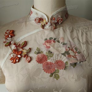 Può includere: Un abito cheongsam beige con colletto alla coreana e bottoni a nodo decorativi. Il tessuto presenta un sottile motivo floreale tono su tono, accentuato da un ricamo floreale nei toni dell'arancione, del rosa e del bianco sul petto.